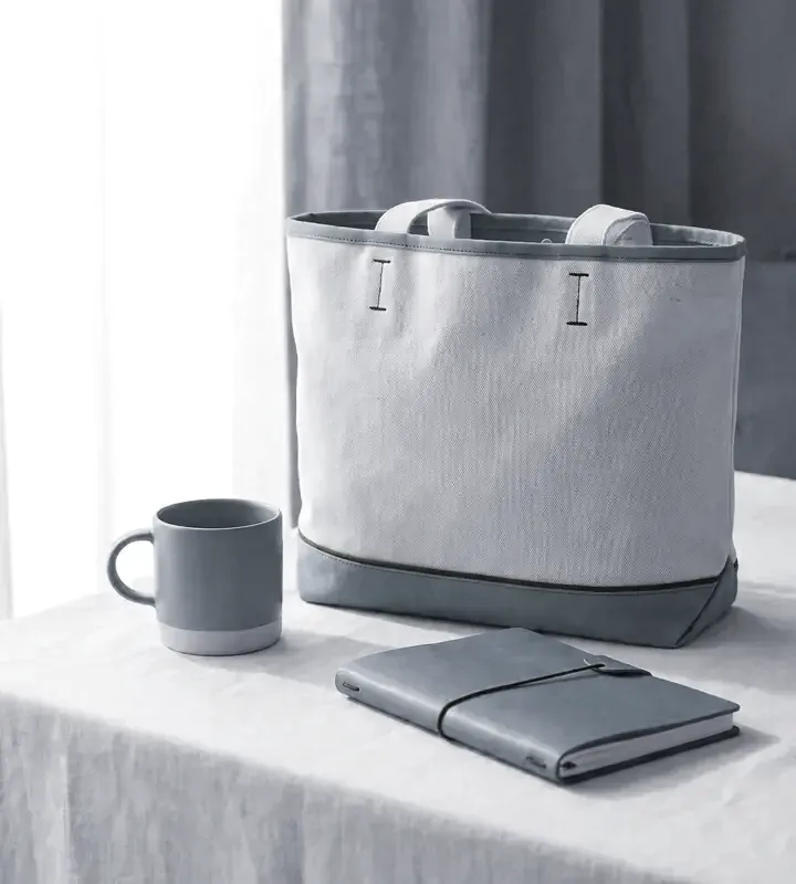 A canvas tote bag with a blue base, a blue mug and a notebook on a linen-covered table
